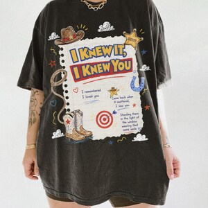 May include: A black t-shirt featuring a graphic print of a notebook page with the text I KNEW IT, I KNEW YOU. The design includes illustrations of a cowboy hat, a sheriff badge, a lasso, cowboy boots, a target, and various small decorative icons.