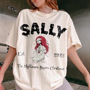 Op de afbeelding: Cr&egrave;mekleurig T-shirt met de naam "SALLY" in zwarte, druipende letters. Daaronder een illustratie van een personage met rood haar en verband, met de tekst "Est 1993 The Nightmare Before Christmas".