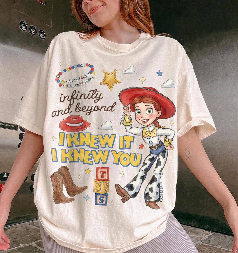 Pode incluir: Uma t-shirt de manga curta cor de creme com uma impress&atilde;o gr&aacute;fica de uma cowgirl com chap&eacute;u vermelho, botas castanhas, um colar de contas e blocos de letras. O texto diz infinity and beyond e I KNEW IT I KNEW YOU.