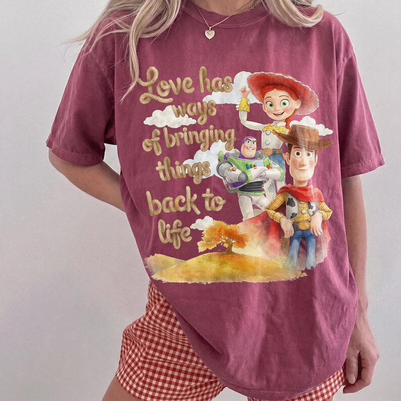Pu&ograve; includere: Una t-shirt a maniche corte color bordeaux con una stampa grafica di Woody, Buzz Lightyear e Jessie di Toy Story. Il testo recita, Love has ways of bringing things back to life, accompagnato da un'illustrazione paesaggistica con un albero e colline.