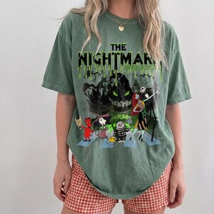 May include: Green t-shirt featuring a graphic from "The Nightmare Before Christmas." The design includes the film's title in dripping green letters, along with characters like Jack Skellington and Sally. The shirt is paired with red and white checkered shorts.