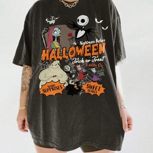 K&ouml;nnte beinhalten: Ein anthrazitfarbenes T-Shirt mit einem Grafikdruck der Charaktere aus Nightmare Before Christmas. Das Design zeigt Jack Skellington, Sally und Oogie Boogie mit dem Text Nightmare Before Halloween Trick or Treat Candy Co.
