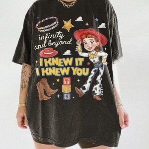 Pode incluir: Uma t-shirt preta de manga curta com uma estampa gr&aacute;fica de uma cowgirl, um chap&eacute;u vermelho, botas de cowboy castanhas e blocos de letras. O texto diz infinity and beyond, I KNEW IT I KNEW YOU e You've got a friend in me numa pulseira de contas.