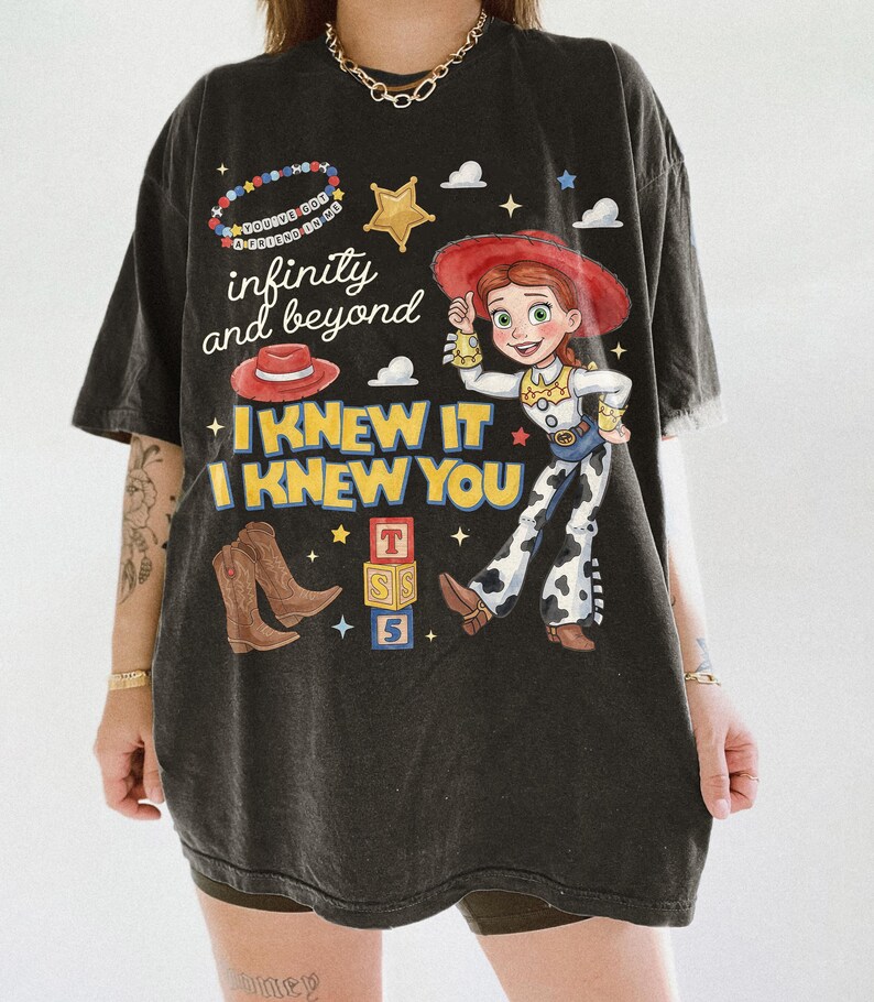 Pode incluir: Uma t-shirt preta de manga curta com uma estampa gr&aacute;fica de uma cowgirl, um chap&eacute;u vermelho, botas de cowboy castanhas e blocos de letras. O texto diz infinity and beyond, I KNEW IT I KNEW YOU e You've got a friend in me numa pulseira de contas.