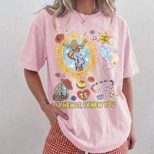 May include: A light pink short-sleeve t-shirt featuring a graphic print with a gold ornate frame, a cartoon character playing guitar, a star with the text INFINITY AND BEYOND, balloons, a ball, and the text I KNEW IT, I KNEW YOU in yellow.