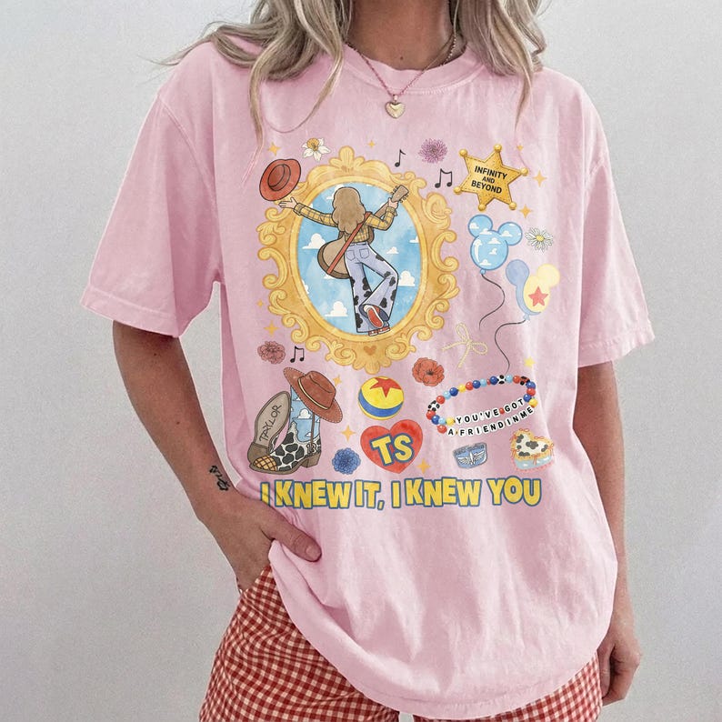 May include: A light pink short-sleeve t-shirt featuring a graphic print with a gold ornate frame, a cartoon character playing guitar, a star with the text INFINITY AND BEYOND, balloons, a ball, and the text I KNEW IT, I KNEW YOU in yellow.