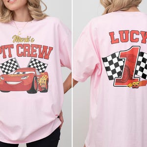 Op de afbeelding: Lichtroze T-shirt met een rode raceauto-afbeelding en de tekst "Mark's Pit Crew" op de voorkant. De achterkant toont de naam "Lucy", het nummer "1" en geblokte vlaggen. Het shirt heeft een relaxte, oversized pasvorm.