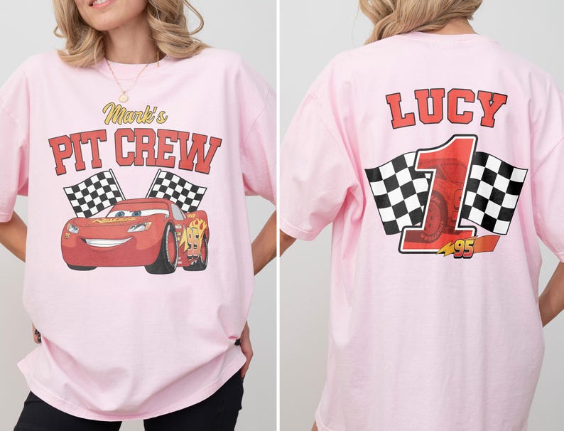 Op de afbeelding: Lichtroze T-shirt met een rode raceauto-afbeelding en de tekst "Mark's Pit Crew" op de voorkant. De achterkant toont de naam "Lucy", het nummer "1" en geblokte vlaggen. Het shirt heeft een relaxte, oversized pasvorm.