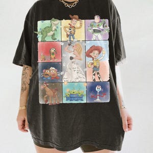K&ouml;nnte beinhalten: Ein schwarzes Kurzarm-T-Shirt mit einem Raster aus farbenfrohen Illustrationen verschiedener Zeichentrickfiguren, darunter ein Dinosaurier, ein Cowboy, ein Weltraum-Ranger, ein Hund, ein Kartoffelpaar, ein Pferd, Au&szlig;erirdische, eine Gabel und eine S&auml;ngerin.