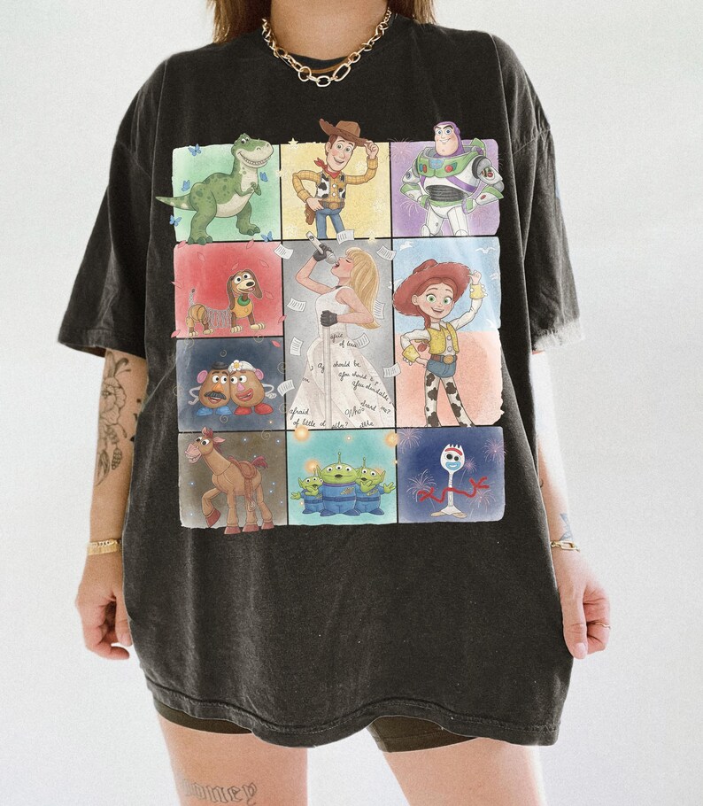 K&ouml;nnte beinhalten: Ein schwarzes Kurzarm-T-Shirt mit einem Raster aus farbenfrohen Illustrationen verschiedener Zeichentrickfiguren, darunter ein Dinosaurier, ein Cowboy, ein Weltraum-Ranger, ein Hund, ein Kartoffelpaar, ein Pferd, Au&szlig;erirdische, eine Gabel und eine S&auml;ngerin.