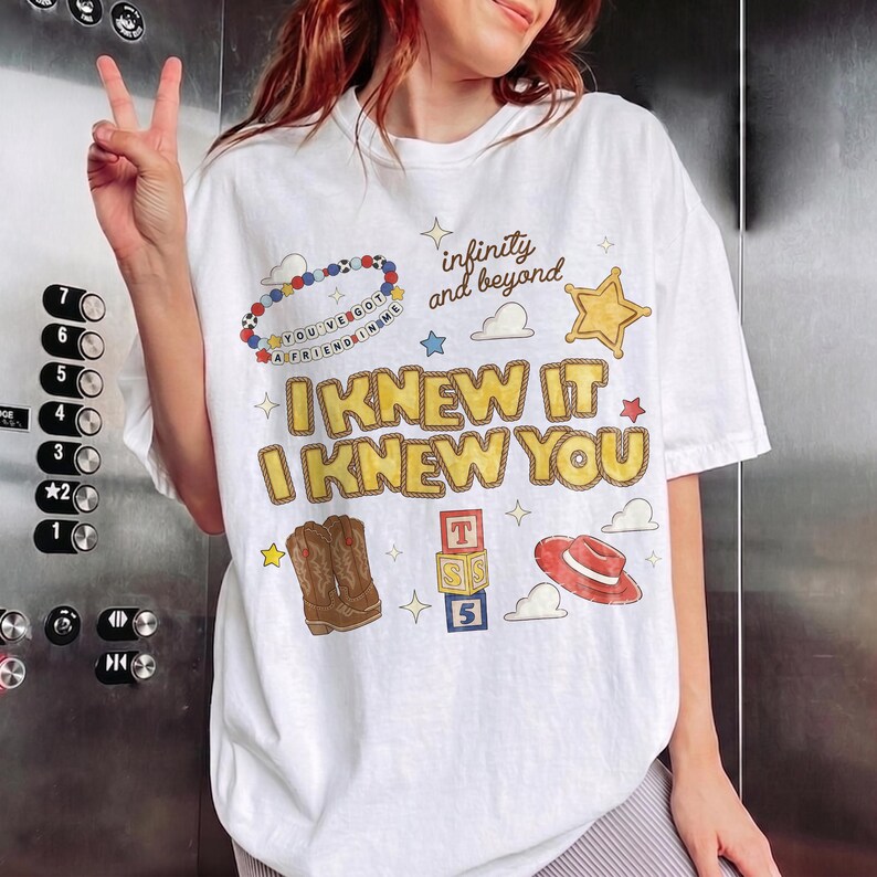 May include: A white short-sleeve t-shirt featuring a graphic print with the text "I KNEW IT I KNEW YOU" in yellow, "infinity and beyond" in brown, and "YOU'VE GOT A FRIEND IN ME" on a beaded circle, alongside illustrations of cowboy boots, a hat, and blocks.