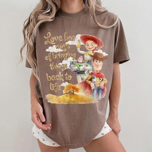 Pu&ograve; includere: Una t-shirt marrone a maniche corte con una stampa grafica di Buzz Lightyear, Jessie e Woody di Toy Story. Il testo Love has ways of bringing things back to life si trova sopra un'illustrazione paesaggistica con un albero e colline.