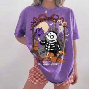 May include: A purple t-shirt featuring a graphic of a skeleton character holding a purple lollipop. The design includes a tombstone with the text NAME 2026, pumpkins, a castle, and the text Nightmare on Main Street in a decorative frame.