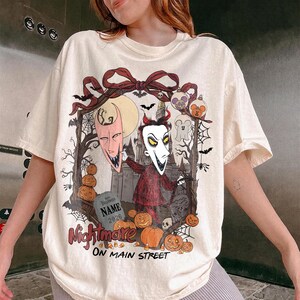 May include: An off-white t-shirt featuring a graphic print of a character with a pointed mask, a red suit, and horns. The design includes a tombstone with the text 'NAME 2026', pumpkins, bats, spiderwebs, and the text 'Nightmare ON MAIN STREET'.