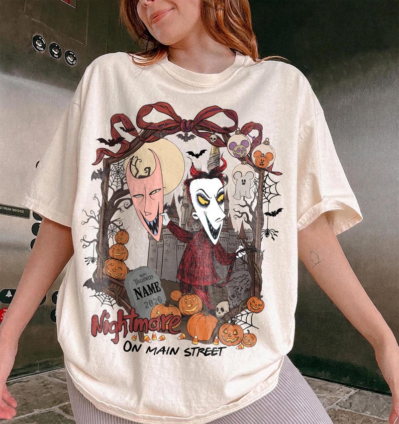 May include: An off-white t-shirt featuring a graphic print of a character with a pointed mask, a red suit, and horns. The design includes a tombstone with the text 'NAME 2026', pumpkins, bats, spiderwebs, and the text 'Nightmare ON MAIN STREET'.
