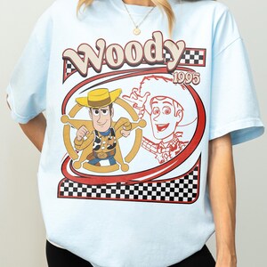 May include: Light blue t-shirt featuring a graphic of the character Woody from the movie Toy Story. The design includes the word "Woody" in a retro font, the year "1995", and a checkered pattern.