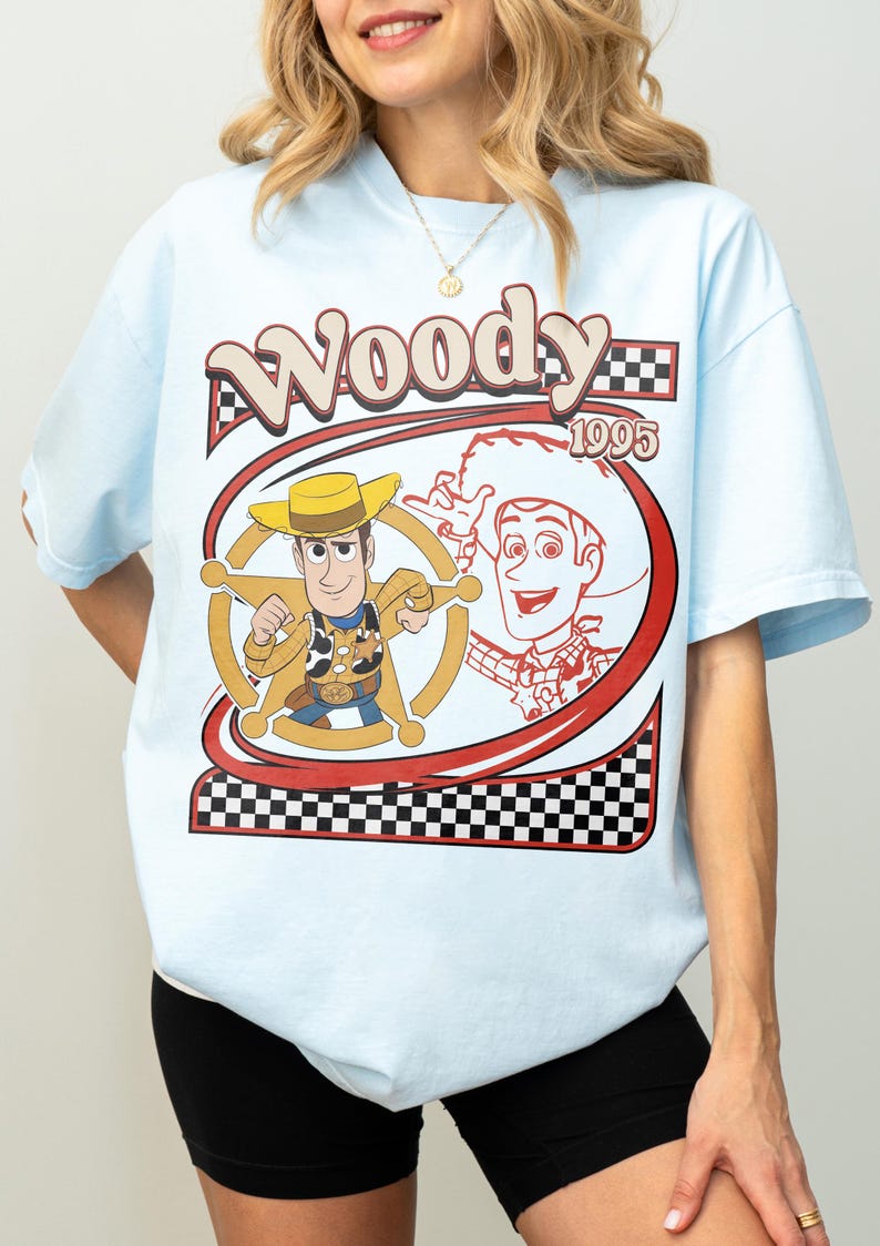 May include: Light blue t-shirt featuring a graphic of the character Woody from the movie Toy Story. The design includes the word "Woody" in a retro font, the year "1995", and a checkered pattern.