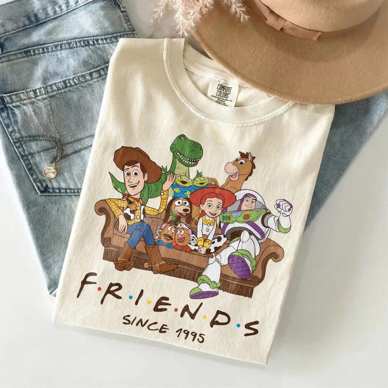 May include: Cream-colored t-shirt featuring a graphic of Toy Story characters on a brown couch. The word "FRIENDS" is printed below the graphic in a colorful font, with "SINCE 1995" underneath. The shirt is folded on top of blue jeans.