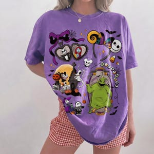 K&ouml;nnte beinhalten: Ein lila T-Shirt mit einem farbenfrohen Grafikdesign mit Charakteren und Elementen aus "The Nightmare Before Christmas". Das Design umfasst Jack Skellington, Sally, Oogie Boogie und andere Halloween-Motive.