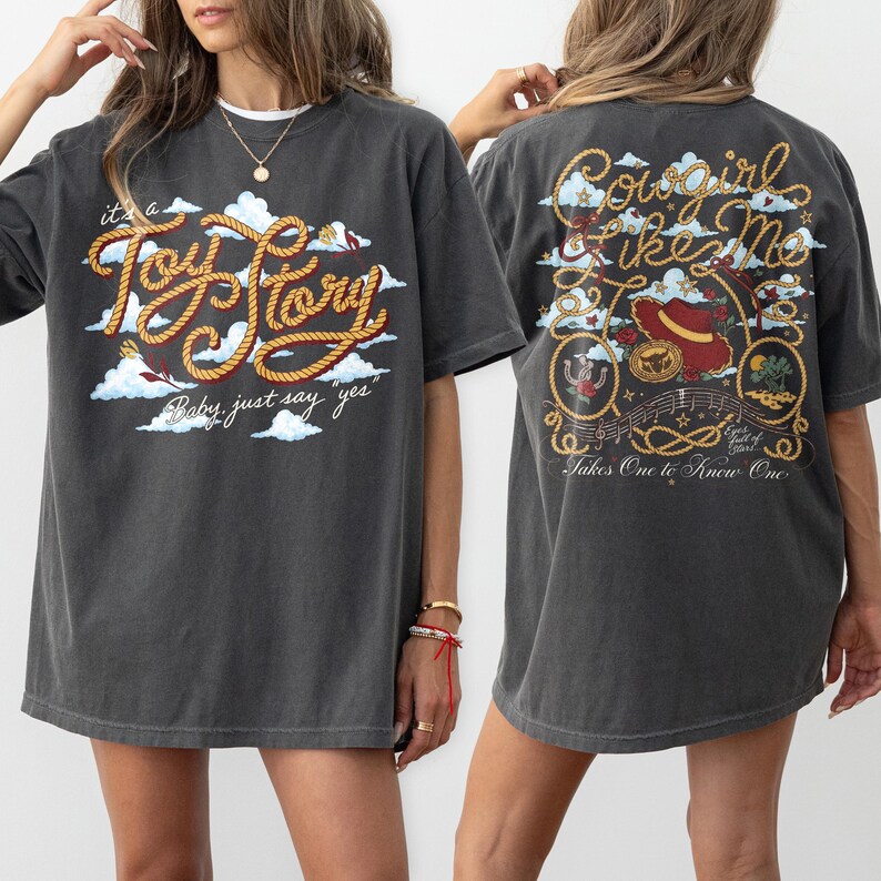 Op de afbeelding: Een antracietgrijs t-shirt met korte mouwen en een vooropdruk met de tekst "it's a Toy Story Baby, just say yes" in touwstijl letters en wolken. De achterkant toont "Cowgirl Like Me" met een cowboyhoed en touwmotieven.