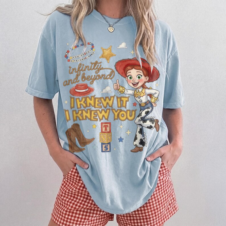 May include: A light blue t-shirt featuring a graphic print of a cowgirl character, a red hat, brown cowboy boots, a yellow star, and building blocks. The text reads "infinity and beyond", "I KNEW IT", "I KNEW YOU", and "YOU'VE GOT A FRIEND IN ME".