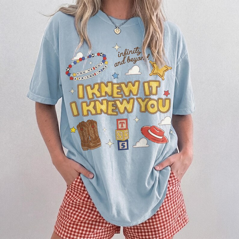 May include: A light blue short-sleeve t-shirt featuring a graphic print with the text "I KNEW IT I KNEW YOU", "infinity and beyond", and "YOU'VE GOT A FRIEND IN ME". The design includes illustrations of cowboy boots, a red hat, and blocks.