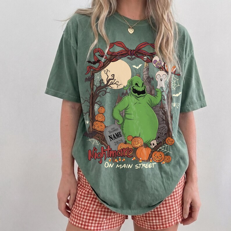 May include: A sage green short-sleeve t-shirt featuring a graphic print of a green character, pumpkins, a tombstone with the text NAME 2026, and a red bow. The text Nightmare ON MAIN STREET is printed in red and white below the central graphic.