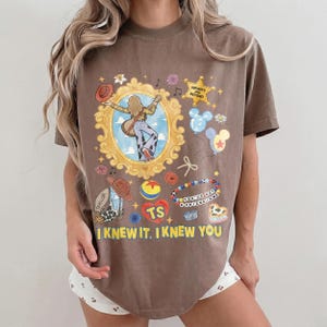 May include: A brown short-sleeve t-shirt featuring a graphic print of a person in a gold frame, a star with the text "INFINITY AND BEYOND," balloons, a cowboy boot, a ball, a beaded bracelet with "YOU'VE GOT A FRIEND IN ME," and "I KNEW IT, I KNEW YOU."