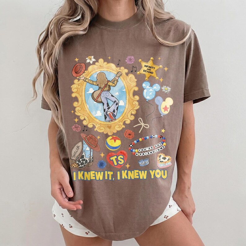 May include: A brown short-sleeve t-shirt featuring a graphic print of a person in a gold frame, a star with the text "INFINITY AND BEYOND," balloons, a cowboy boot, a ball, a beaded bracelet with "YOU'VE GOT A FRIEND IN ME," and "I KNEW IT, I KNEW YOU."