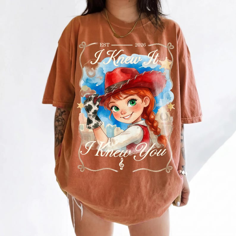 May include: A rust-colored t-shirt featuring a graphic of a cartoon cowgirl with red hair, a red cowboy hat, and a cow-print glove. The text reads EST 2026, I Knew It, and I Knew You, framed by a rope design with stars and a musical note symbol.