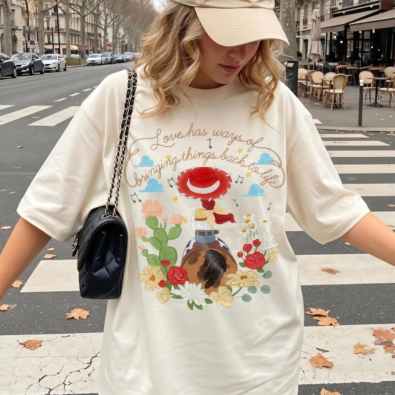 May include: An oversized cream-colored t-shirt featuring a graphic print of a character in a red hat riding a horse, surrounded by cacti and flowers. The text reads, Love has ways of bringing things back to life, accompanied by musical notes.