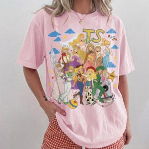 May include: A light pink short-sleeve t-shirt featuring a large graphic print of various animated characters in colorful outfits. The design includes the letters TS, a rocket, stars, clouds, and a sheriff badge. The characters are depicted in a collage style.