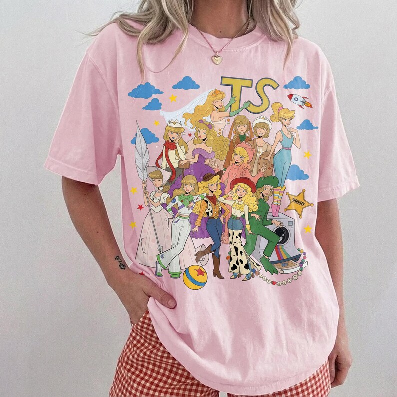 May include: A light pink short-sleeve t-shirt featuring a large graphic print of various animated characters in colorful outfits. The design includes the letters TS, a rocket, stars, clouds, and a sheriff badge. The characters are depicted in a collage style.