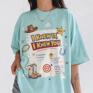 May include: A light blue t-shirt featuring a graphic print of a lined notebook page with the text I KNEW IT, I KNEW YOU. The design includes illustrations of a cowboy hat, a sheriff badge, a lasso, cowboy boots, a horseshoe, and a target.