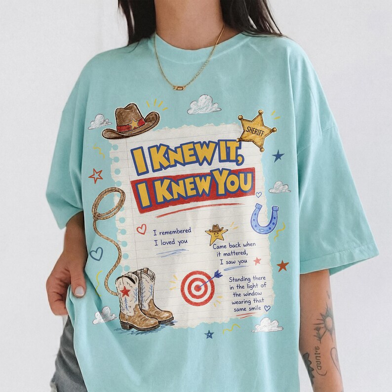 May include: A light blue t-shirt featuring a graphic print of a lined notebook page with the text I KNEW IT, I KNEW YOU. The design includes illustrations of a cowboy hat, a sheriff badge, a lasso, cowboy boots, a horseshoe, and a target.