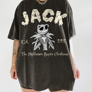 Op de afbeelding: Zwart T-shirt met een afbeelding van Jack Skellington uit "The Nightmare Before Christmas". Het woord "JACK" is gedrukt in een druipend lettertype boven het personage. De tekst "Est. 1993" en "The Nightmare Before Christmas" staan ook op het shirt.