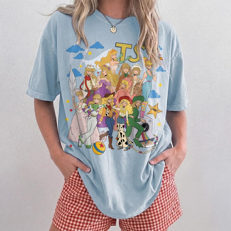 May include: A light blue short-sleeve t-shirt featuring a colorful graphic print of various animated characters in a collage style. The design includes multiple figures, a yellow star, a blue ball with a red star, and a camera illustration.