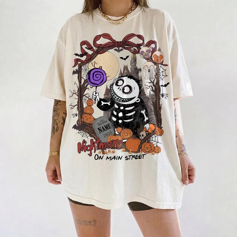 May include: An off-white short-sleeve t-shirt featuring a graphic of a skeleton character holding a purple lollipop. The design includes pumpkins, a tombstone with the text NAME 2026, and the text Nightmare ON MAIN STREET in red and black.