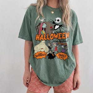 K&ouml;nnte beinhalten: Ein salbeigr&uuml;nes T-Shirt mit einem Grafikdruck der Charaktere aus Nightmare Before Christmas. Der Text lautet Nightmare Before Halloween, Trick or Treat Candy Co., Sugary Surprises und Sweet Scares in orangefarbener und schwarzer Schrift.