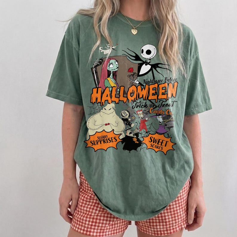 K&ouml;nnte beinhalten: Ein salbeigr&uuml;nes T-Shirt mit einem Grafikdruck der Charaktere aus Nightmare Before Christmas. Der Text lautet Nightmare Before Halloween, Trick or Treat Candy Co., Sugary Surprises und Sweet Scares in orangefarbener und schwarzer Schrift.