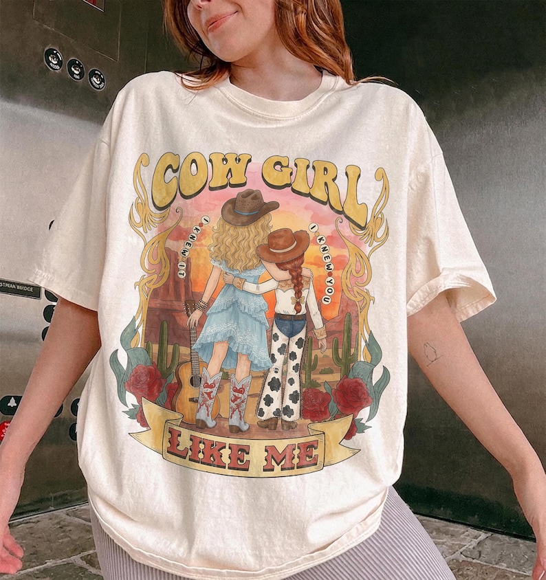 May include: A cream-colored t-shirt featuring a graphic print of two figures in cowboy hats standing in a desert landscape. The text reads COW GIRL and LIKE ME, with the phrase I KNEW IT I KNEW YOU encircling the figures. Red roses and cacti frame the scene.