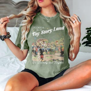 May include: Olive green t-shirt with a graphic of Toy Story Land in Florida. The design includes Mickey Mouse, Woody, Buzz Lightyear, and other characters. Text reads "FLORIDA Est. 2018 Toy Story Land To Infinity and Beyond."