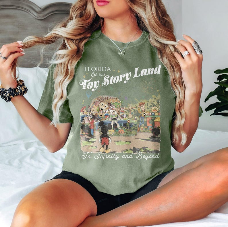 May include: Olive green t-shirt with a graphic of Toy Story Land in Florida. The design includes Mickey Mouse, Woody, Buzz Lightyear, and other characters. Text reads "FLORIDA Est. 2018 Toy Story Land To Infinity and Beyond."