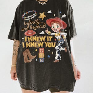 May include: A black short-sleeve t-shirt featuring a graphic of a cowgirl character, a red hat, brown cowboy boots, and alphabet blocks. The text reads "infinity and beyond", "YOU'VE GOT A FRIEND IN ME", and "I KNEW IT I KNEW YOU" in yellow and white.