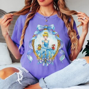 May include: A lilac-colored t-shirt featuring a framed illustration of Cinderella in a blue gown, with two mice. The frame is gold with blue ribbons and bows. The shirt is a comfortable, casual style.