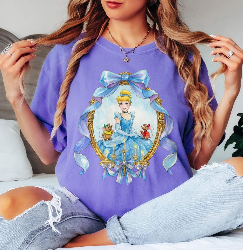 May include: A lilac-colored t-shirt featuring a framed illustration of Cinderella in a blue gown, with two mice. The frame is gold with blue ribbons and bows. The shirt is a comfortable, casual style.