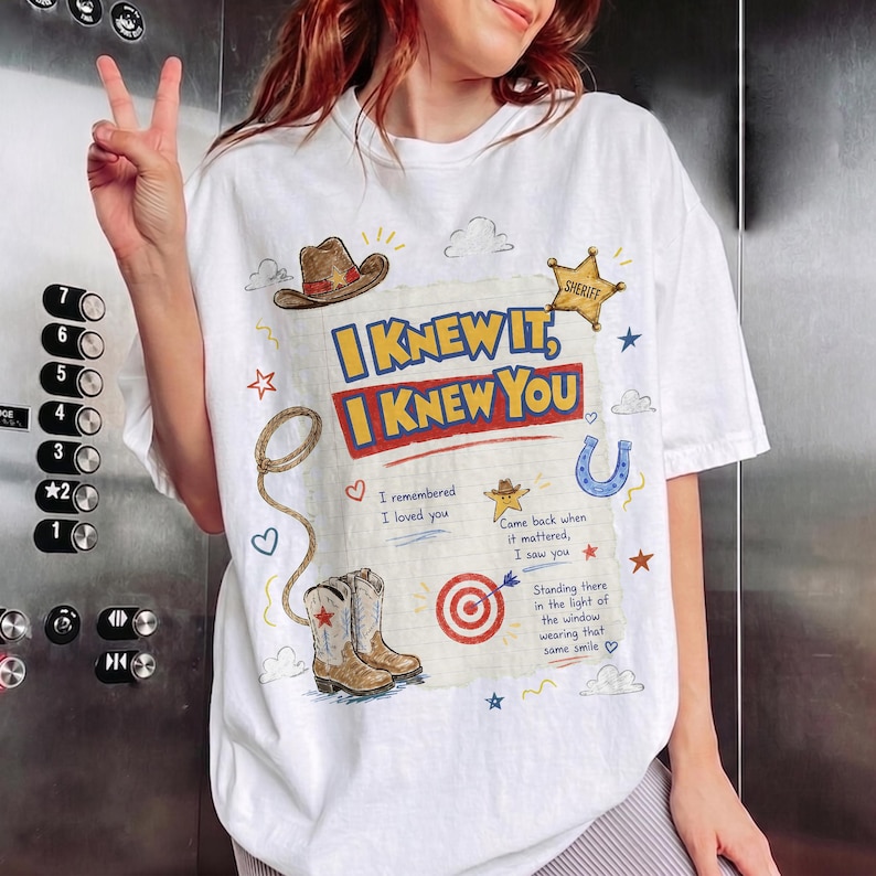 May include: A white t-shirt featuring a graphic print with the text I KNEW IT, I KNEW YOU, along with illustrations of a cowboy hat, a sheriff star, a lasso, a horseshoe, a target, and cowboy boots, plus additional handwritten-style text.