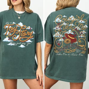 Op de afbeelding: Een donkergroen t-shirt met een voorzijde voorzien van touwachtige tekst "it's a Toy Story, baby, just say yes" tussen wolken. De achterzijde toont "Cowgirl Like Me" en "Takes One to Know One" met een touw, een hoed en sterren.