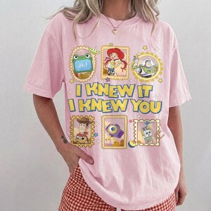 May include: A light pink short-sleeve t-shirt featuring a graphic print of Toy Story characters in postage stamp frames. The text reads I KNEW IT I KNEW YOU in yellow letters with blue outlines. The design includes Rex, Jessie, Buzz Lightyear, Woody, and other icons.
