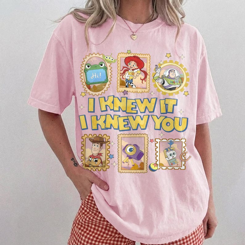 May include: A light pink short-sleeve t-shirt featuring a graphic print of Toy Story characters in postage stamp frames. The text reads I KNEW IT I KNEW YOU in yellow letters with blue outlines. The design includes Rex, Jessie, Buzz Lightyear, Woody, and other icons.